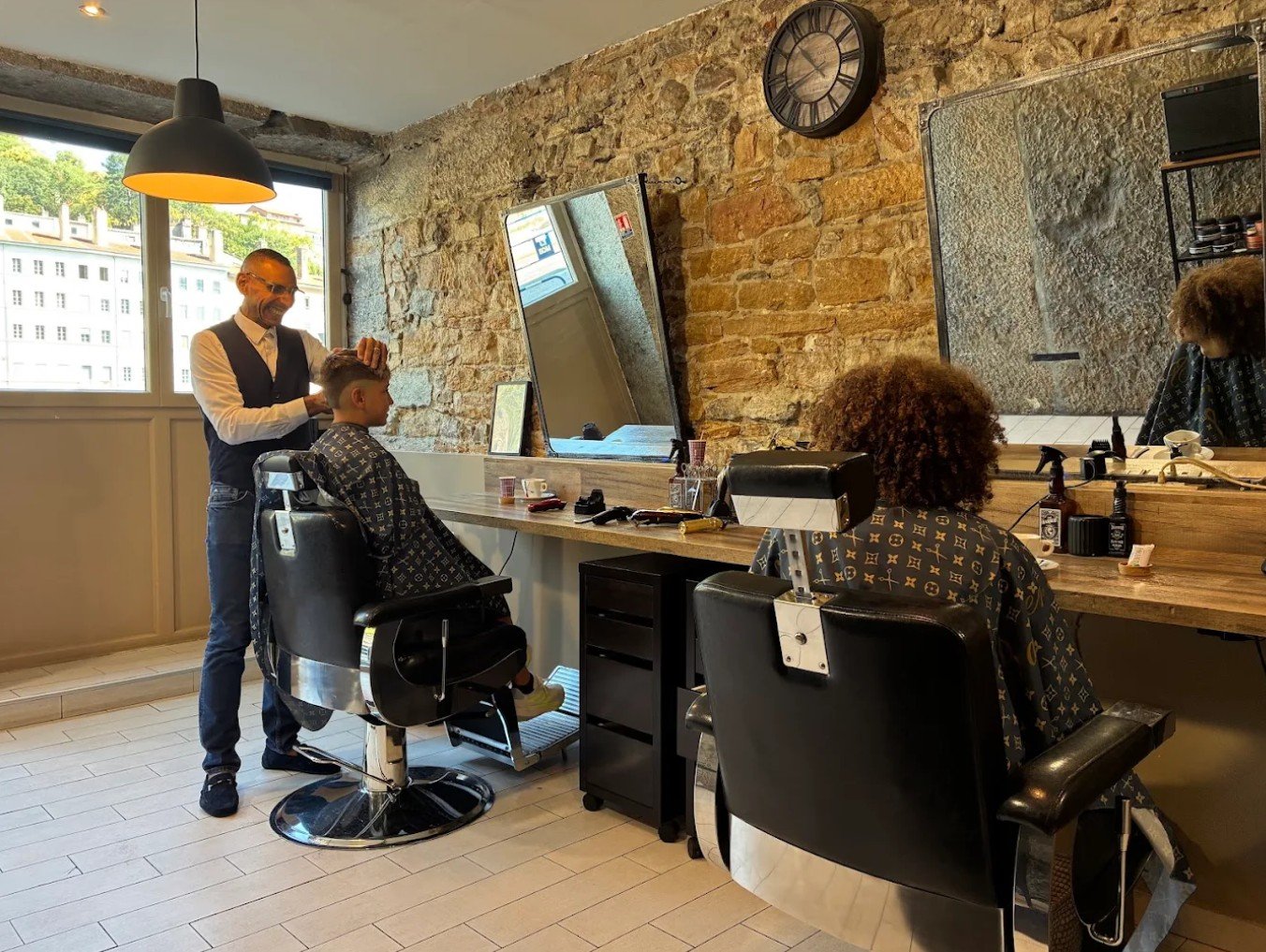 Wahib coupe les cheveux d’un jeune client devant le mur en pierre du salon
